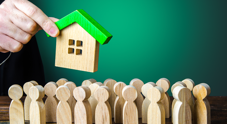 A hand holding a small wooden house above a crowd of wooden figures, symbolizing the high demand for homes and the competitive housing market in Lake Oswego, Oregon.