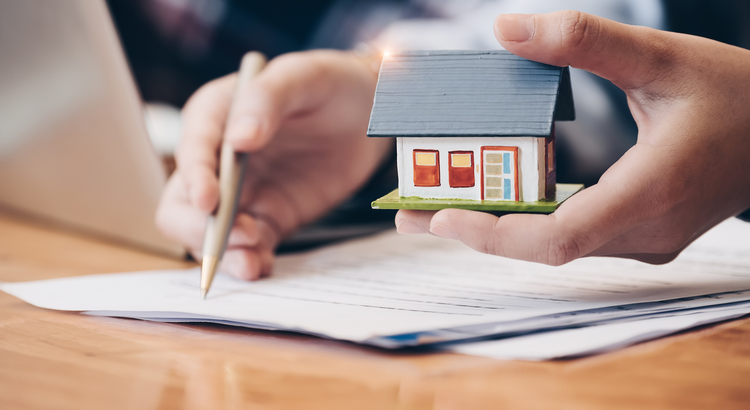 Close-up of hands holding a small house model and signing documents, symbolizing home buying and mortgage agreements.